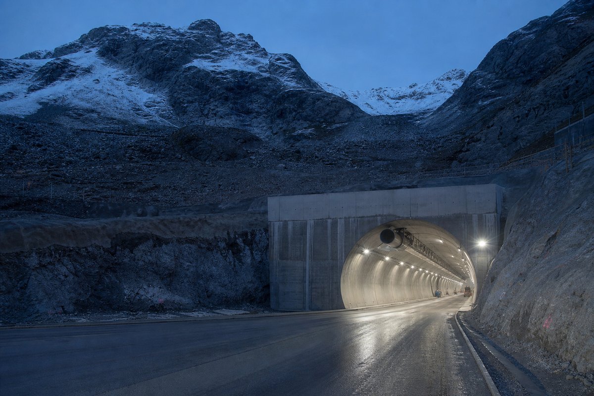 Transport infrastructure structure in Switzerland: high alpine valley, snow on the crags above a massive base-tunnel portal, at blue hour with the portal lit, shot wide at 24mm. (AI-generated image)