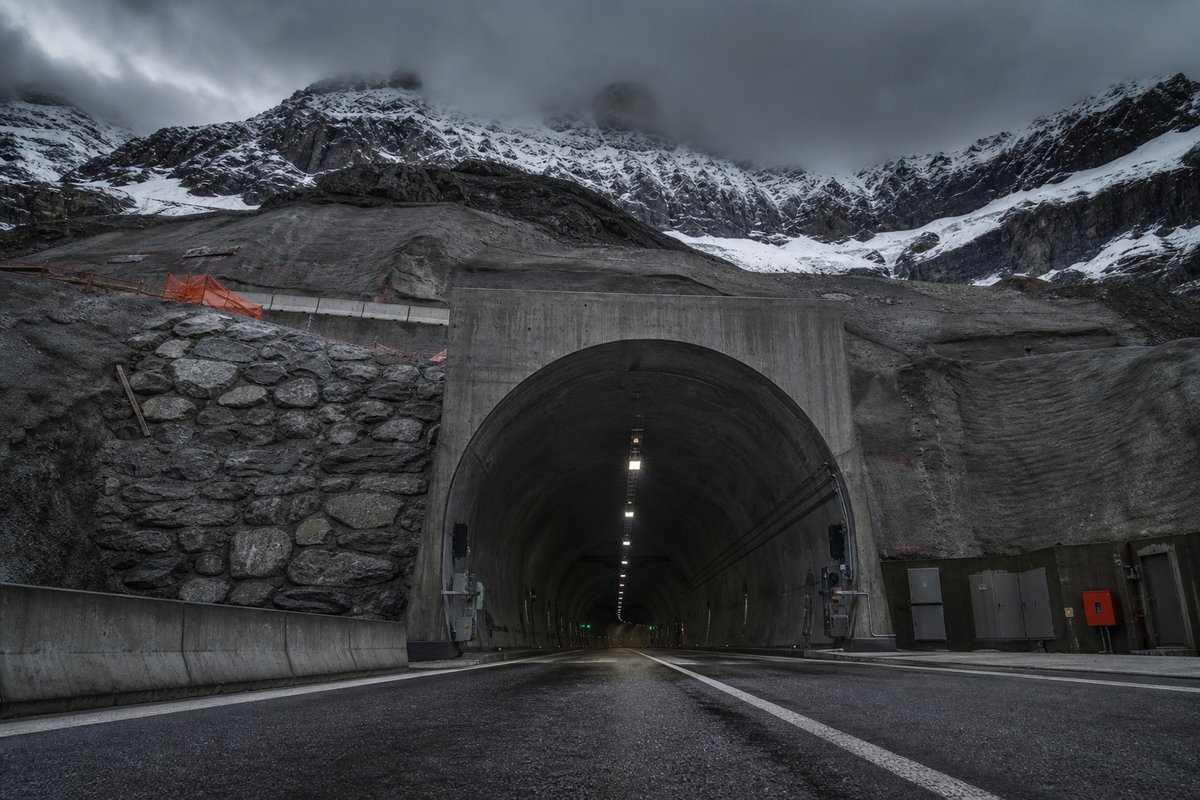 Road tunnel portal in Switzerland: high alpine valley, snow on the crags above a massive base-tunnel portal, under a heavy dark sky, a low camera position near the road surface. (AI-generated image)