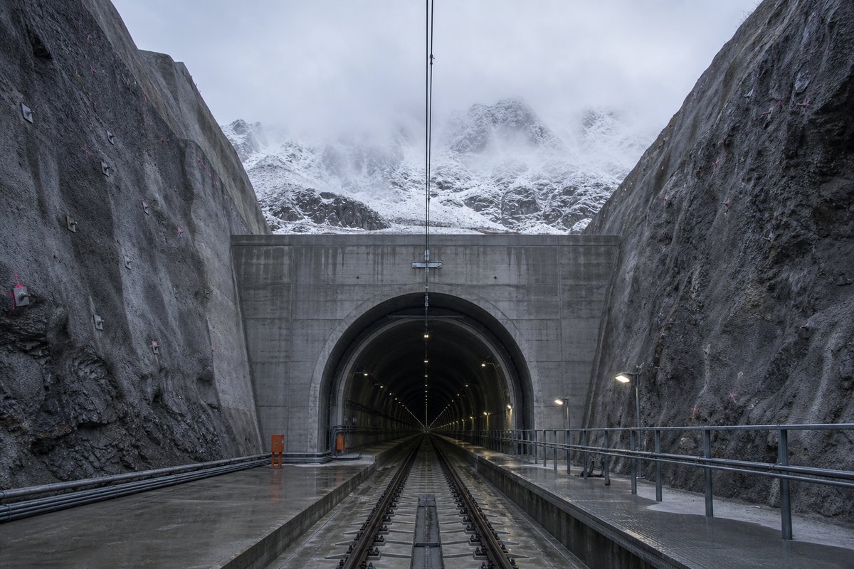 Railway tunnel portal with track running in in Switzerland: high alpine valley, snow on the crags above a massive base-tunnel portal, in thin morning fog. (AI-generated image)