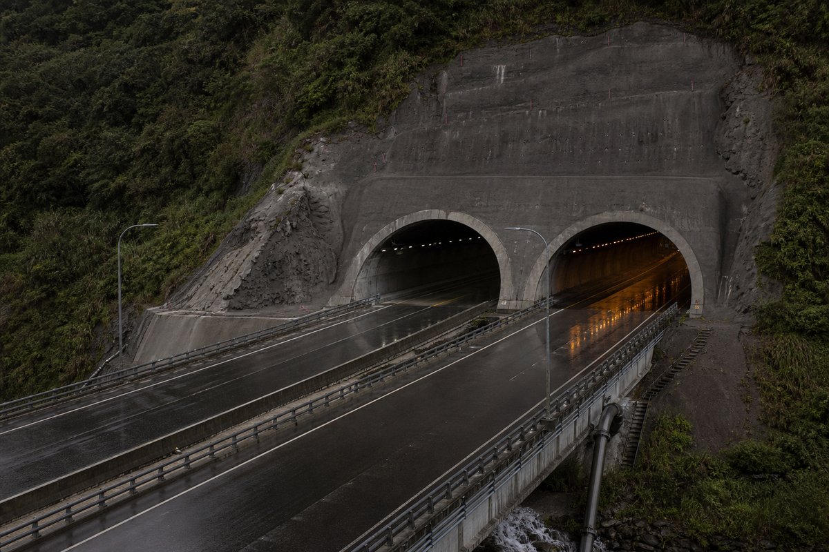 Road tunnel portal in Taiwan: subtropical forested mountain with a broad expressway portal in rain, in hard low winter sun, a slightly elevated three-quarter view. (AI-generated image)