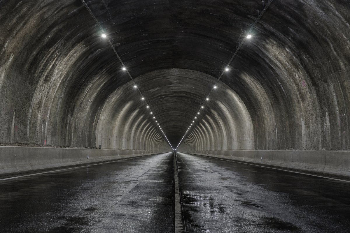 Looking down the axis of an extremely long two-lane road tunnel bore, luminaires receding to a point: a mountainous transport corridor, at dusk with lights just on, shot at 50mm. (AI-generated image)
