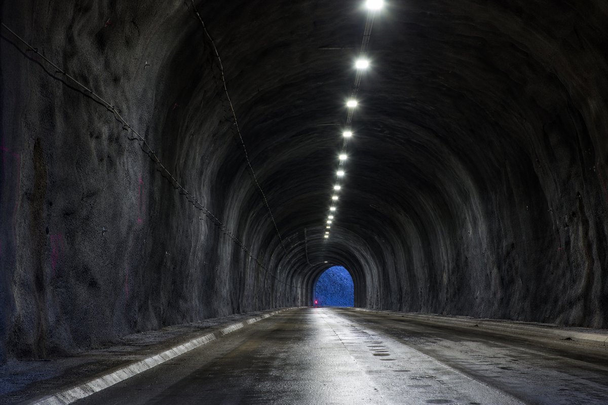 Looking down the axis of an extremely long two-lane road tunnel bore, luminaires receding to a point: a mountainous transport corridor, at blue hour with the portal lit. (AI-generated image)