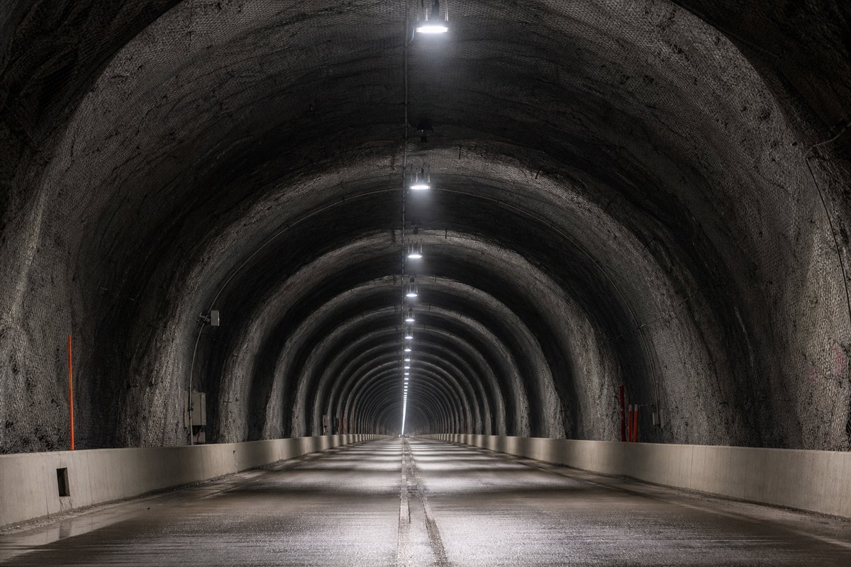Looking down the axis of an extremely long two-lane road tunnel bore, luminaires receding to a point: a mountainous transport corridor, at dusk with lights just on. (AI-generated image)