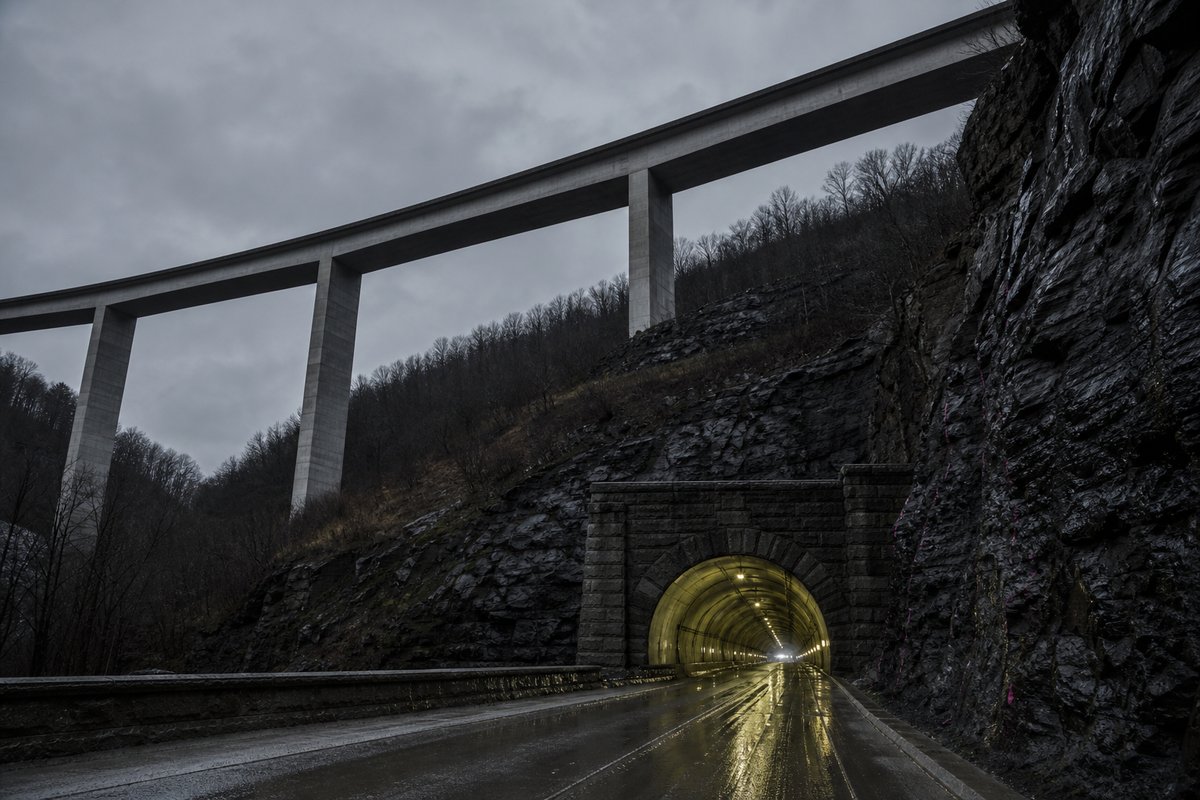 Long concrete box-girder viaduct on tall piers, seen from below in the United States: a mountainous transport corridor, after rain, everything wet and reflective. (AI-generated image)