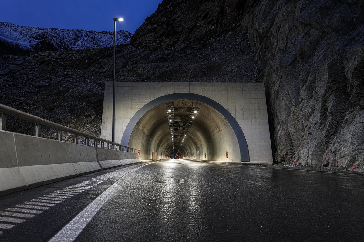 Monumental tunnel portal at dusk, the road vanishing into the mountain: a mountainous transport corridor, at blue hour with the portal lit. (AI-generated image)