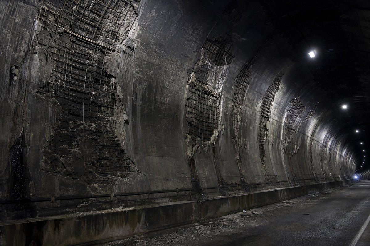 Soot-blackened concrete tunnel lining under emergency lighting, fire-damaged surface spalled away to expose reinforcement, empty carriageway, cold and forensic. (AI-generated image)