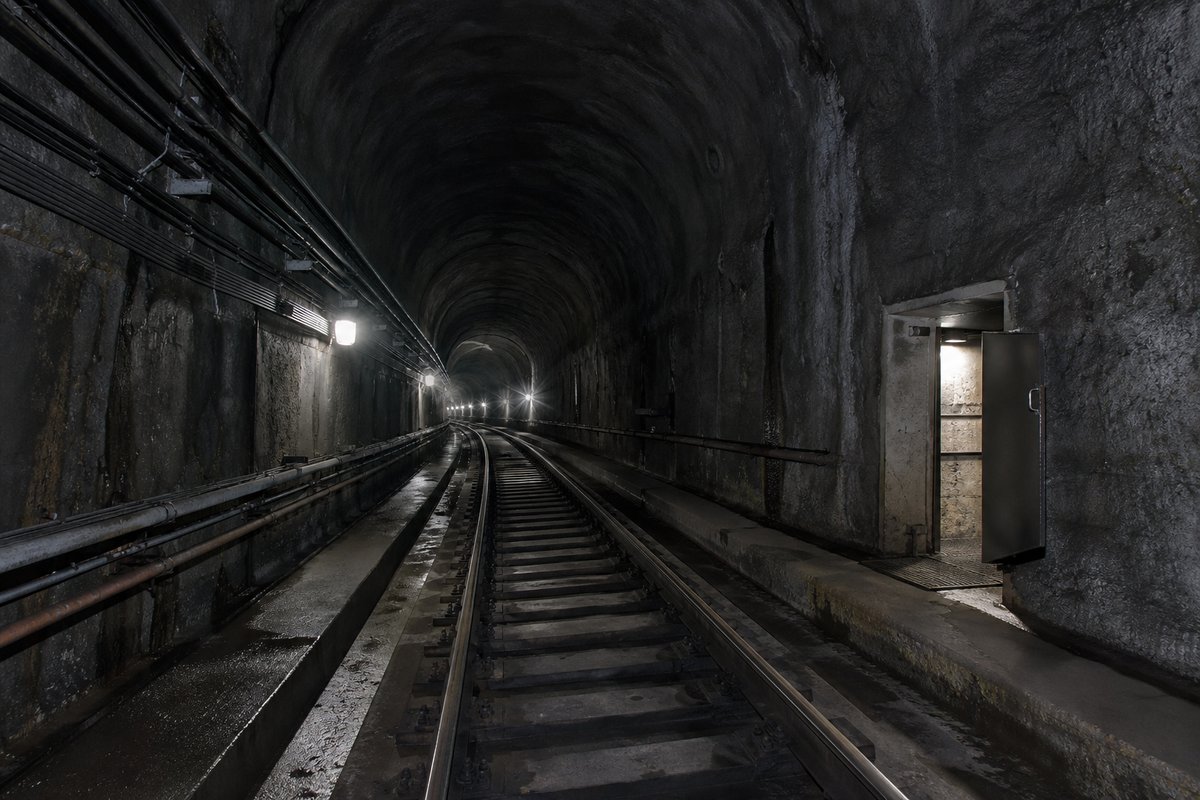 Empty metro tunnel with track and cable runs, dim emergency lighting and a distant lit cross-passage door standing open. (AI-generated image)