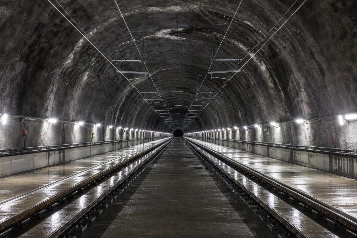 Double-track railway tunnel interior with ballastless slab track and catenary wires receding into darkness, after rain, everything wet and reflective. (immagine generata con AI)