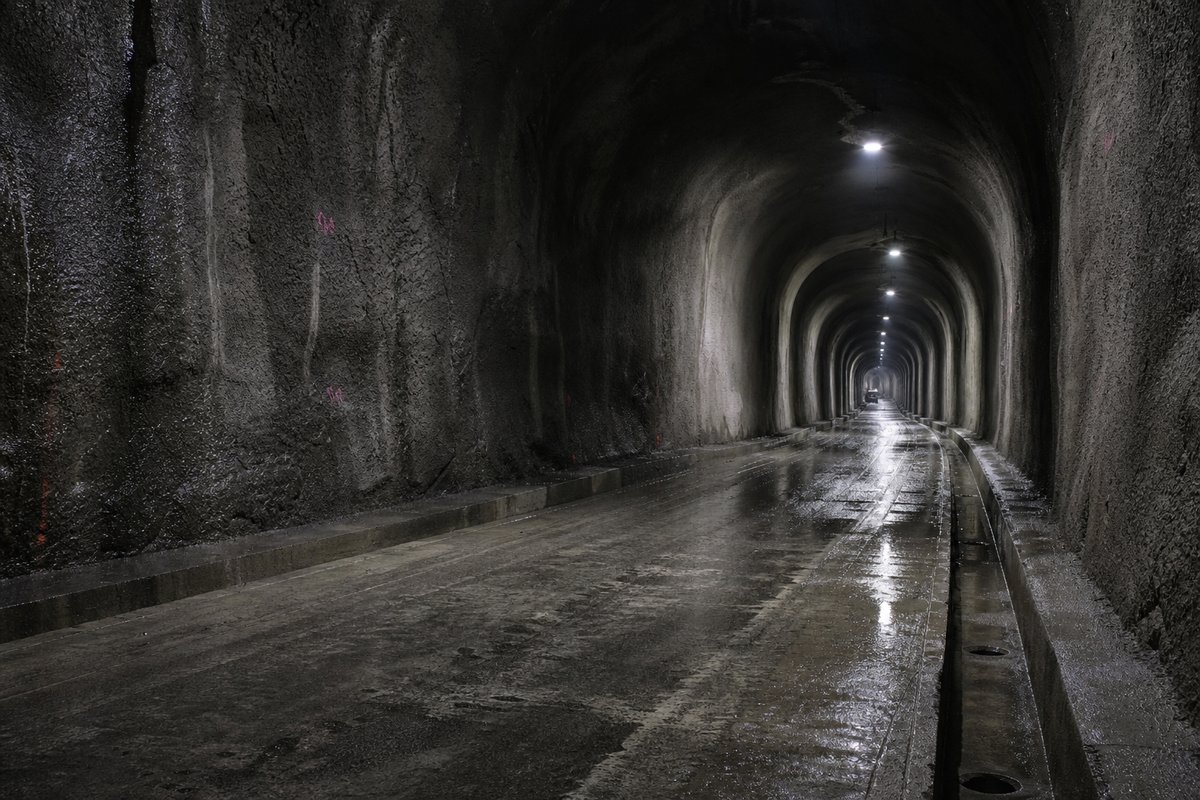 Road tunnel descending steeply below sea level, wet shotcrete walls and deep drainage channels, after rain, everything wet and reflective, shot at 35mm. (immagine generata con AI)