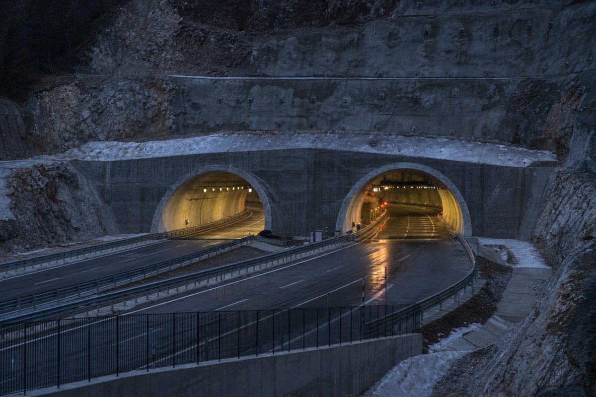 Monumental twin-portal Italian autostrada tunnel mouth in an Apennine limestone cutting at dusk, both bores lit, in cold clear light, shot at 85mm from a distance. (immagine generata con AI)