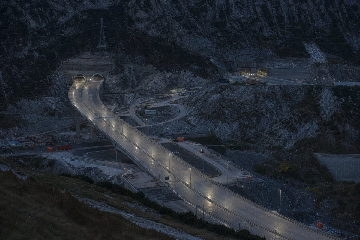 Wide establishing shot of a mountain transport corridor: a mountainous transport corridor, at dusk with lights just on, shot at 50mm, compressed. (AI-generated image)