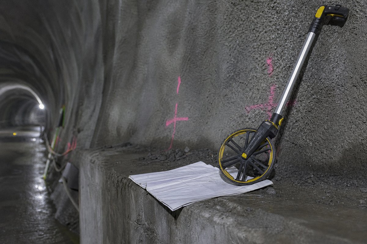 Surveyor's measuring wheel and a folded drawing on a concrete ledge inside a tunnel bore, chainage markings painted on the shotcrete wall behind. (AI-generated image)
