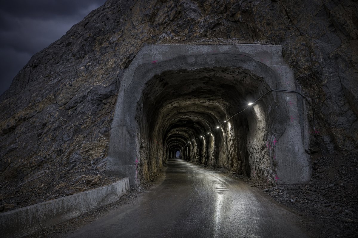 Old hand-drilled rock tunnel with an irregular unlined profile, single lane, weathered portal, under a heavy dark sky, shot at 85mm from a distance. (bilete generert med AI)