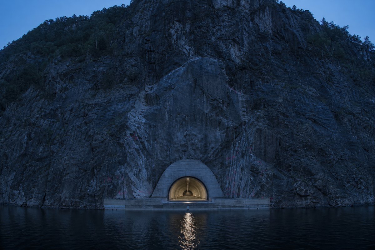 Norwegian fjord crossing seen from above: a tunnel portal at the waterline beneath a steep mountainside, at blue hour with the portal lit, a dead-centre symmetrical composition. (bilete generert med AI)