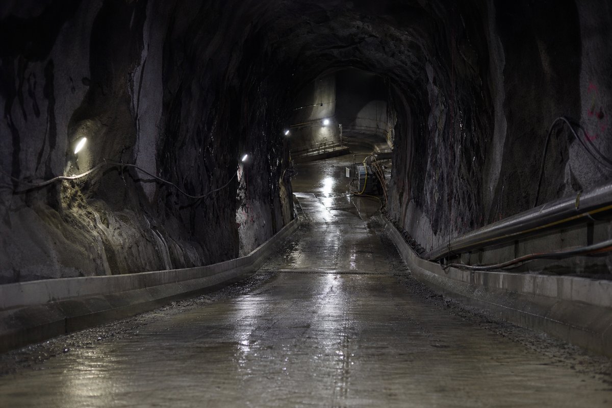 Steep descending bore of a Norwegian subsea tunnel, wet walls, pumping sump and gradient markings, after rain, everything wet and reflective, shot at 50mm, compressed. (bilete generert med AI)