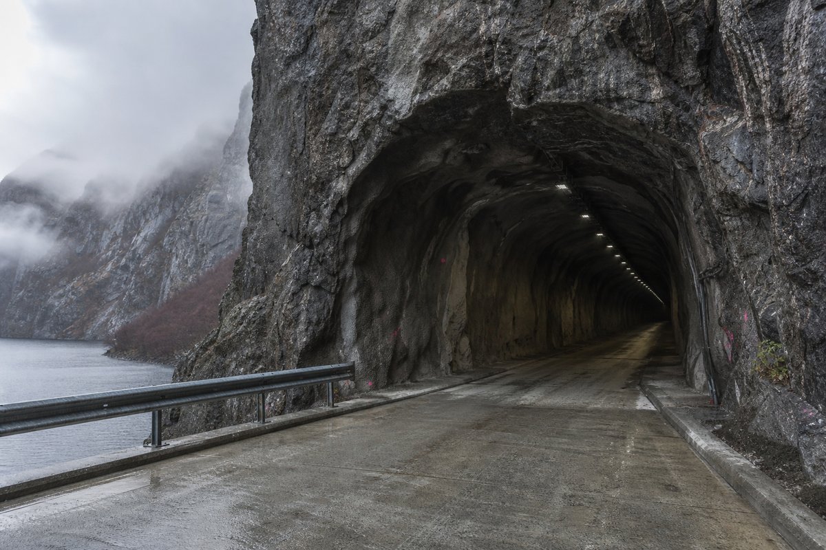 Undecorated blasted rock tunnel mouth on a Norwegian fjord road, wet granite and guardrail, in drifting mist, shot at 35mm. (bilete generert med AI)