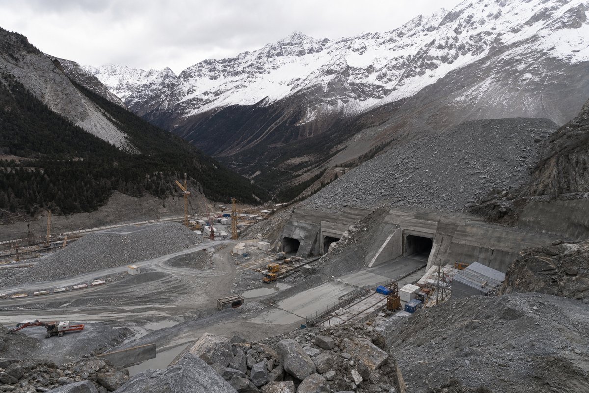 Alpine valley construction site beneath high snow-covered peaks, railway base-tunnel portal works and spoil heaps, overcast. (AI-generated image)