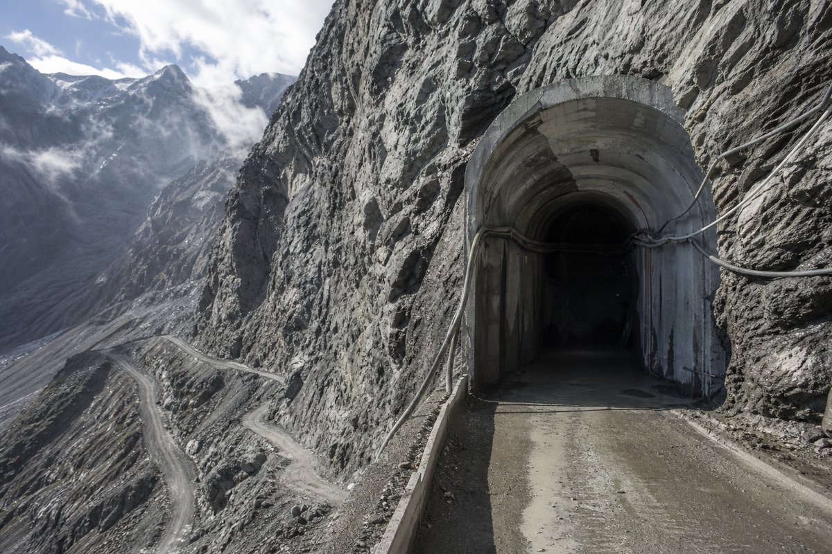 High Alpine railway construction adit driven into a bare rock massif, access road switchbacks below, hard low sun. (AI-generated image)