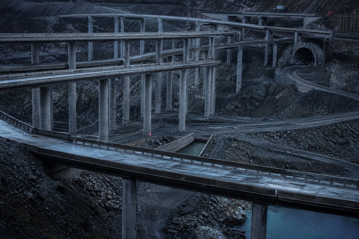 Vast interchange of stacked concrete viaducts and a tunnel portal seen together from a high vantage point, infrastructure layered on infrastructure, at blue hour. (AI-generated image)