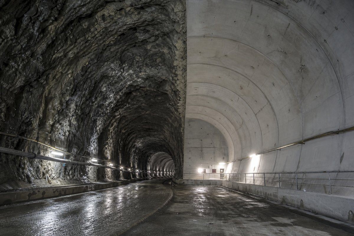 Split-condition tunnel heading: rough blasted rock on one side, a smooth machine-bored segmental lining on the other, meeting at a junction. (AI-generated image)