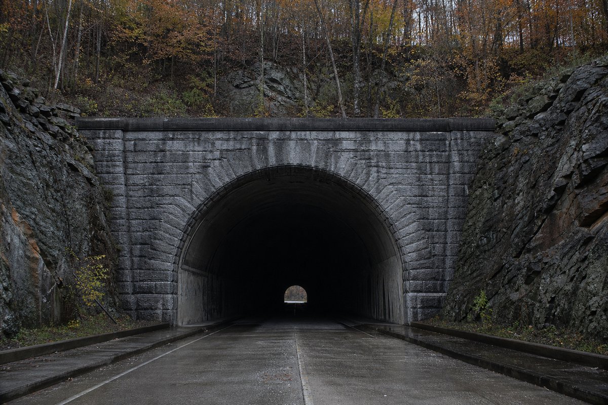 1930s stone-arch highway tunnel portal in an Appalachian rock cut, autumn forest above, flat grey light. (AI-generated image)